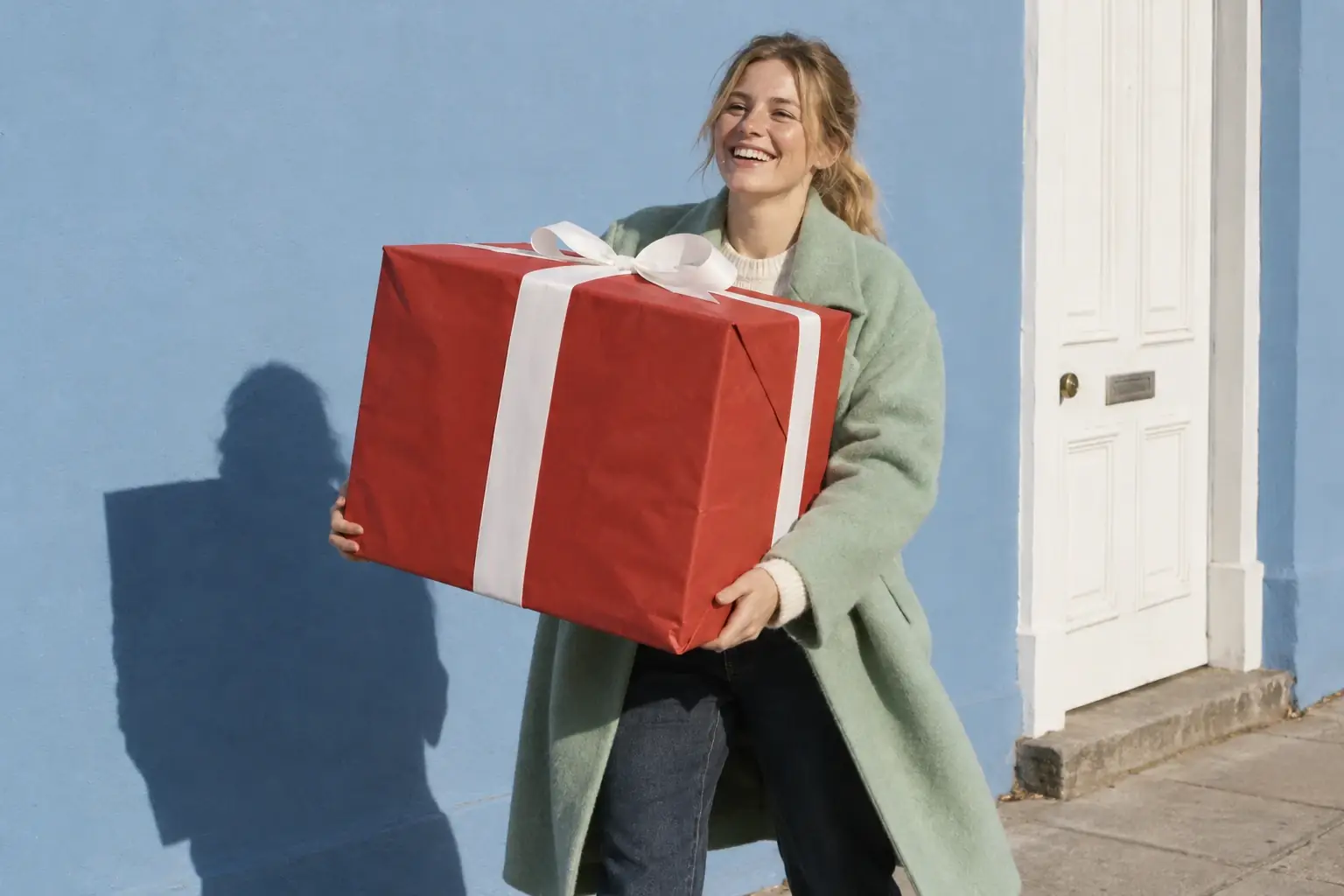 A young blonde woman in a mint green coat walks down the street laughing while carrying a giant gift box wrapped in red paper, in front of a sunny blue wall