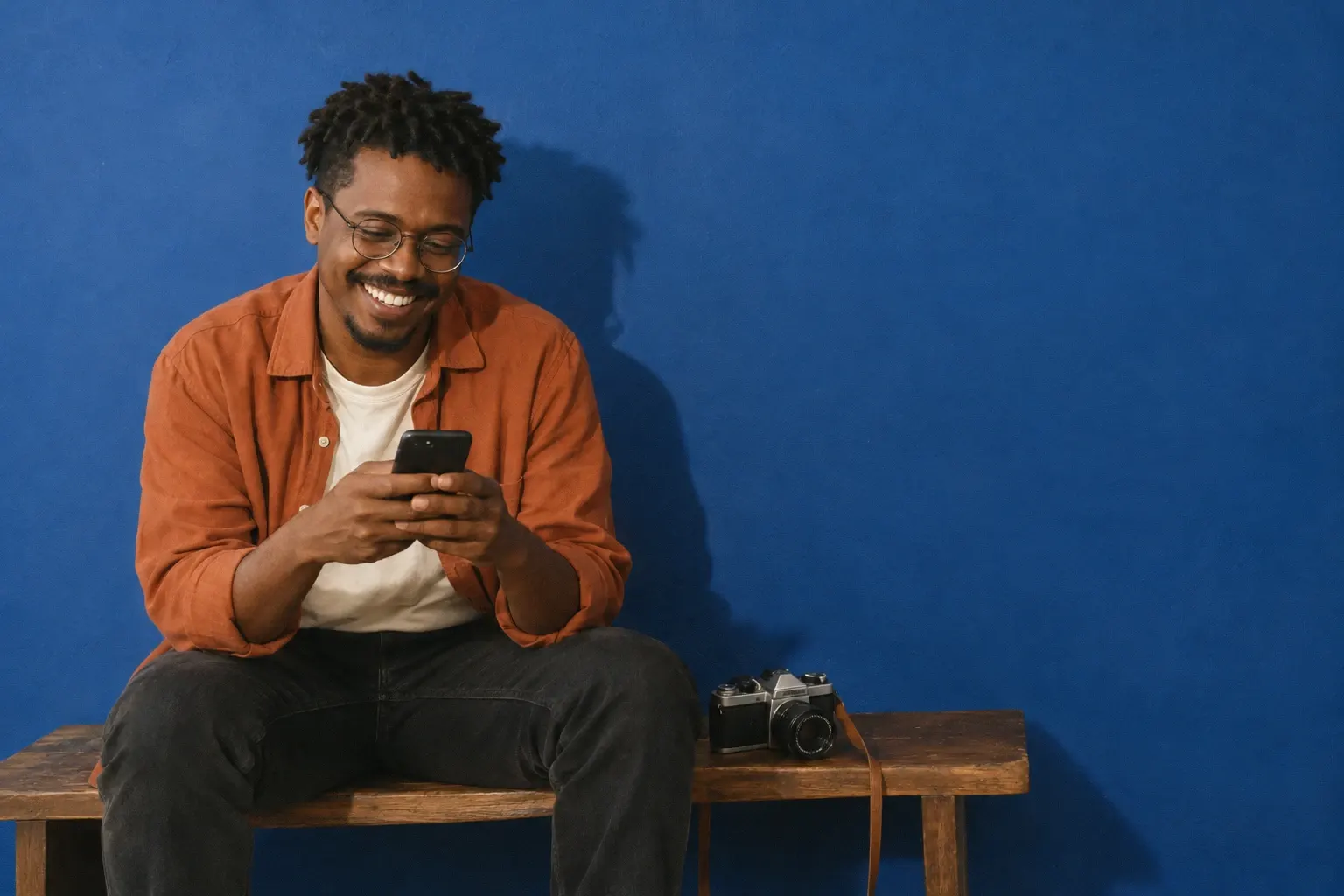 Un photographe de mariage brésilien d’une trentaine d’années, aux twists courts et lunettes rondes, surchemise couleur rouille et t-shirt crème, rit en regardant son téléphone, assis sur un banc en bois devant un mur bleu cobalt, un appareil photo argentique à côté de lui.