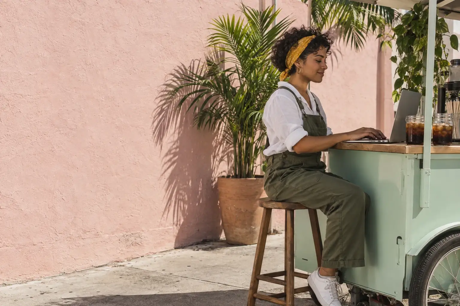 Une femme portoricaine d’environ vingt-huit ans, cheveux bouclés attachés dans un bandana jaune, salopette vert olive et chemise blanche en lin, tape sur son ordinateur portable à côté de son chariot de café glacé vert menthe, devant un mur rose au soleil à Miami.