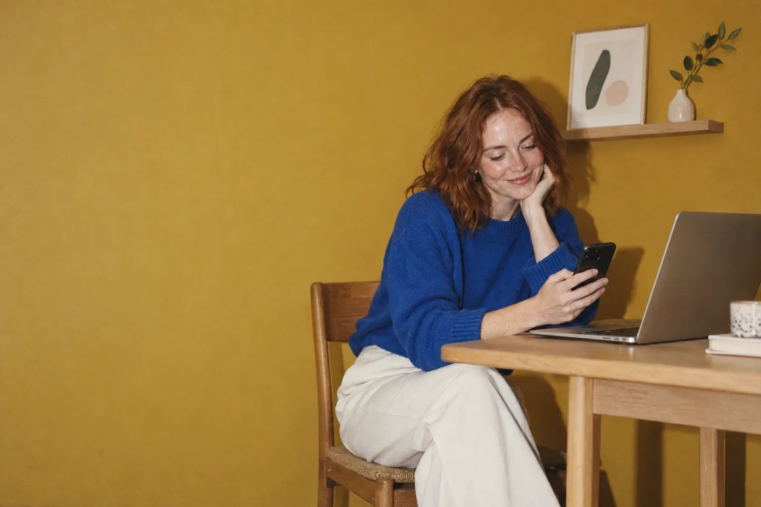 An American content creator in her late twenties, with copper red hair and freckles, in a cobalt blue sweater, smiles at her phone next to her laptop, sitting in front of a mustard yellow wall with a small framed print on a shelf.
