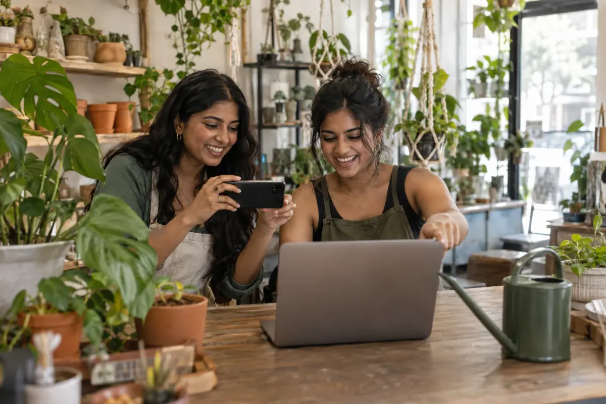 Deux jeunes sœurs au comptoir de leur boutique de plantes : l’une filme avec son téléphone l’écran de l’ordinateur et l’autre le montre du doigt en riant, entourées de pots