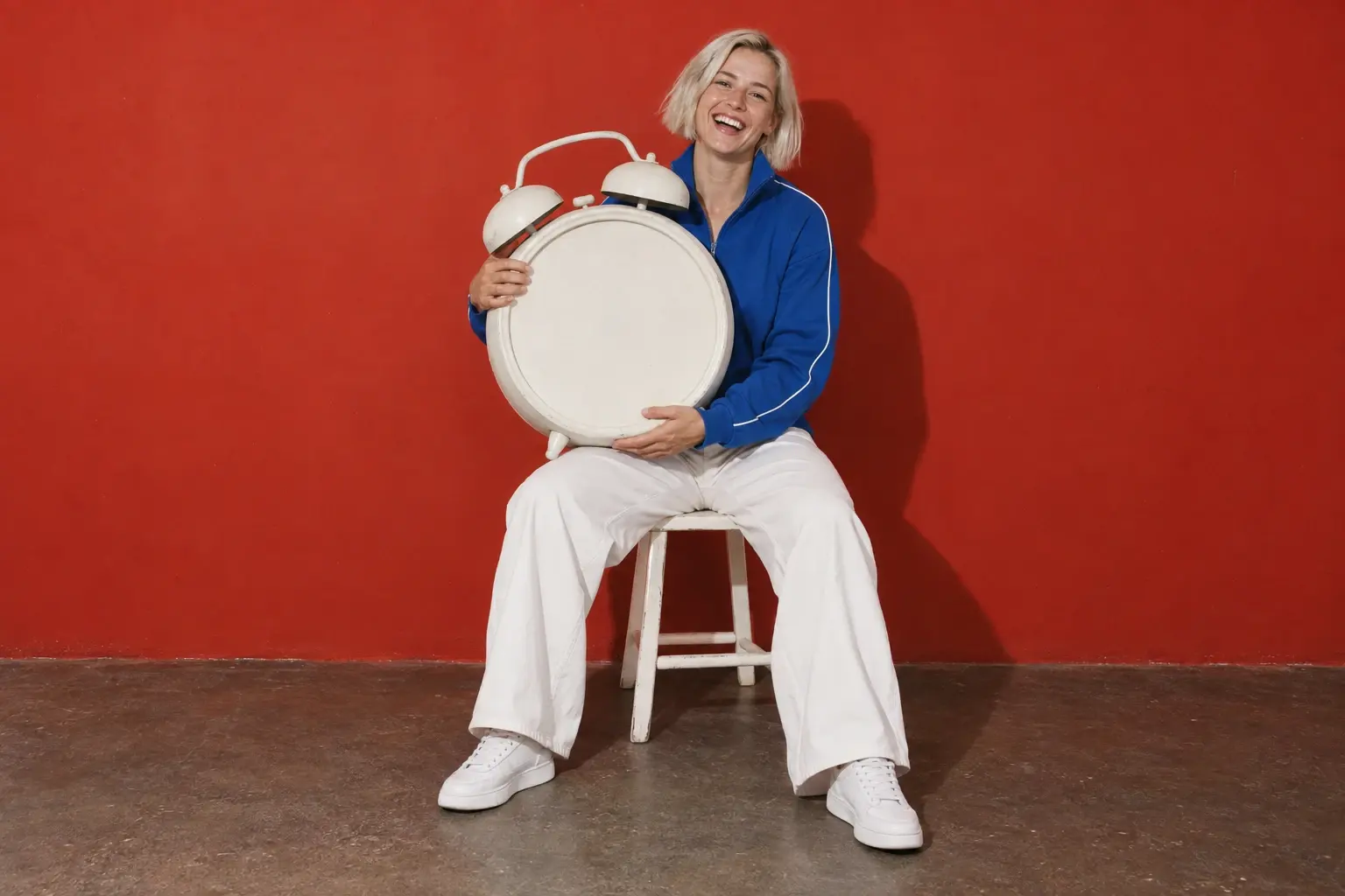 Une coach sportive et créatrice de contenu, aux cheveux blond platine courts et en veste de sport bleu cobalt, rit en serrant un réveil blanc géant, assise sur un tabouret devant un mur rouge